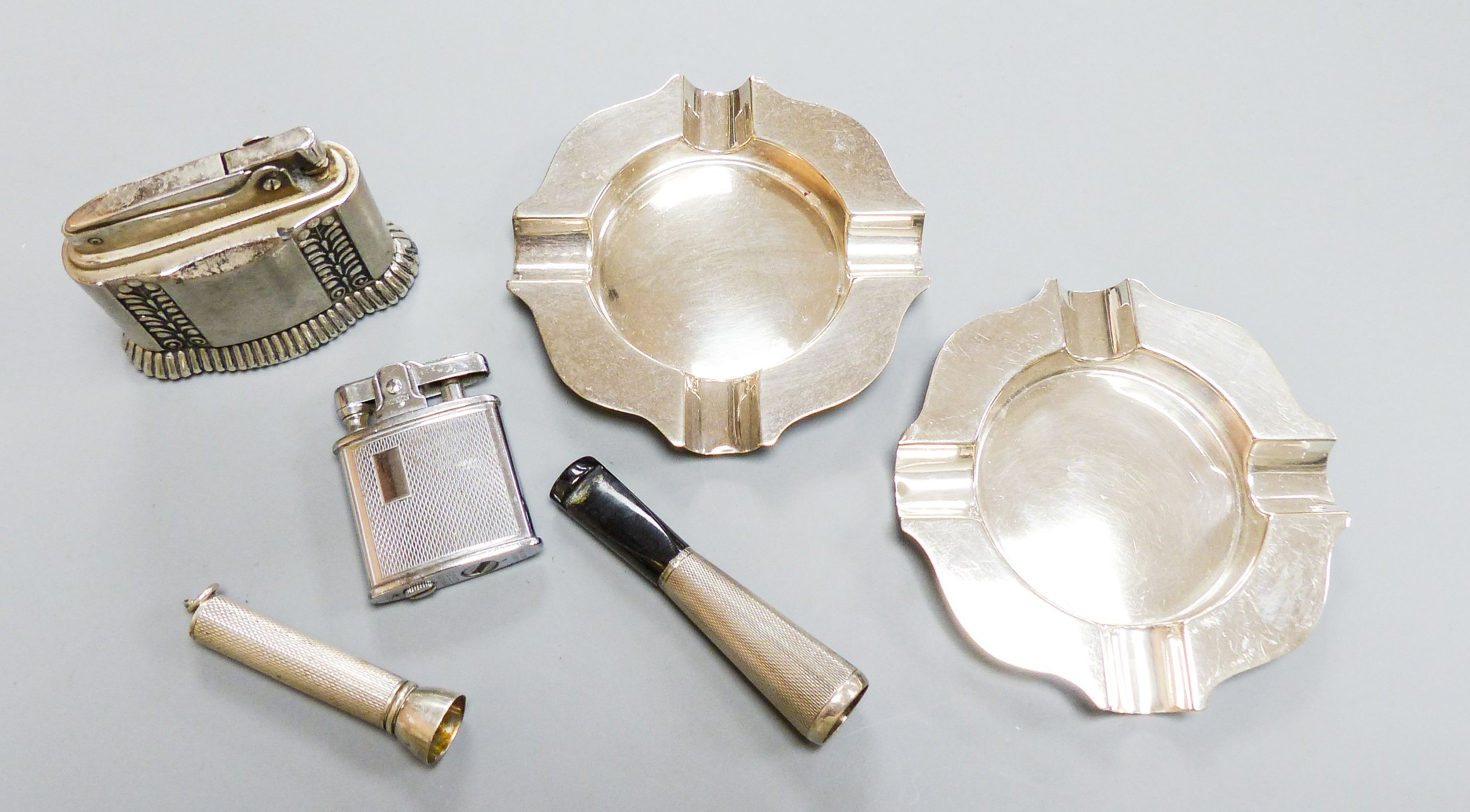 A pair of modern silver ashtrays, two metal lighters, a silver mounted cigar piercer and a white metal cigarette holder.